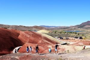 Group of people hiking on a colorful, red and yellow hilly terrain with a lake and mountains in the background under a clear blue sky in Painted Hills Oregon