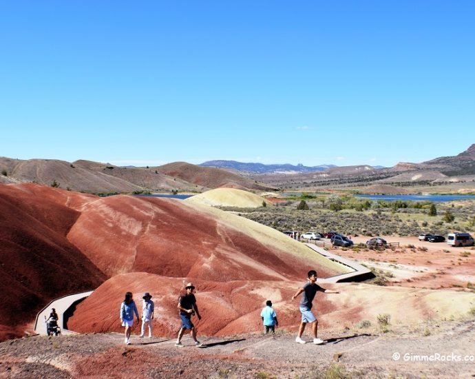 Group of people hiking on a colorful, red and yellow hilly terrain with a lake and mountains in the background under a clear blue sky in Painted Hills Oregon